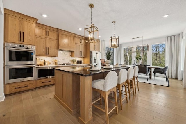 kitchen with pendant lighting, custom exhaust hood, light hardwood / wood-style flooring, an island with sink, and appliances with stainless steel finishes