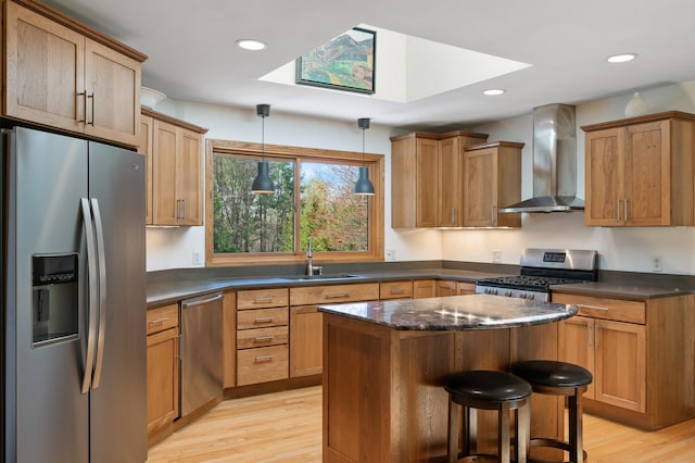 kitchen with wall chimney range hood, a kitchen island, a sink, stainless steel appliances, and light wood-style flooring