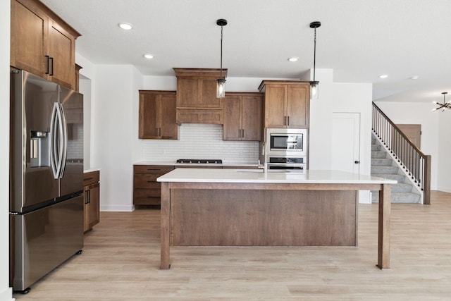 kitchen featuring backsplash, decorative light fixtures, a kitchen island with sink, appliances with stainless steel finishes, and light wood-type flooring