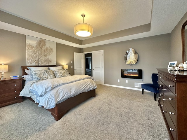 carpeted bedroom featuring a textured ceiling, a tray ceiling, a glass covered fireplace, and baseboards