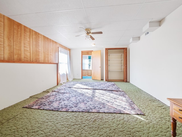 carpeted empty room with a drop ceiling, wood walls, and ceiling fan