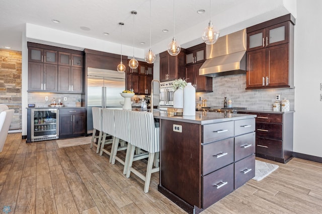 kitchen featuring appliances with stainless steel finishes, wall chimney exhaust hood, a kitchen island with sink, light hardwood / wood-style flooring, and wine cooler