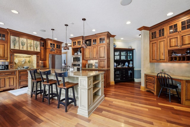 kitchen with light stone counters, decorative light fixtures, a kitchen island with sink, appliances with stainless steel finishes, and light wood-type flooring