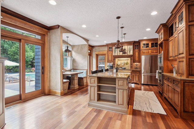 kitchen with appliances with stainless steel finishes, light stone counters, pendant lighting, a center island with sink, and light hardwood / wood-style floors