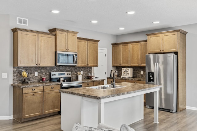 kitchen with a textured ceiling, stainless steel appliances, sink, light hardwood / wood-style flooring, and an island with sink