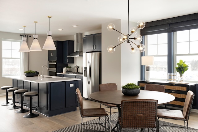 kitchen featuring wall chimney exhaust hood, stainless steel appliances, an island with sink, pendant lighting, and light hardwood / wood-style floors