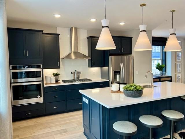 kitchen featuring hanging light fixtures, stainless steel appliances, wall chimney range hood, an island with sink, and a kitchen bar