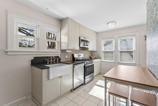 kitchen featuring dark countertops, decorative backsplash, appliances with stainless steel finishes, light tile patterned flooring, and a sink