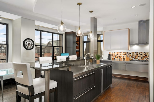 kitchen featuring sink, hanging light fixtures, wall chimney range hood, an island with sink, and a breakfast bar