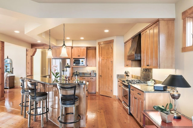 kitchen featuring dark stone counters, wall chimney range hood, pendant lighting, an island with sink, and stainless steel appliances