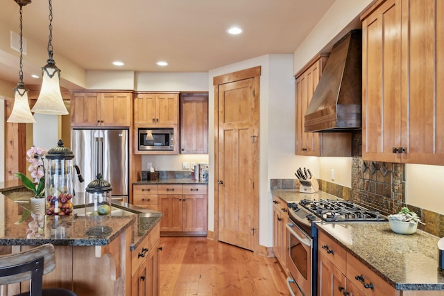 kitchen featuring decorative light fixtures, wall chimney range hood, stainless steel appliances, dark stone counters, and light hardwood / wood-style floors