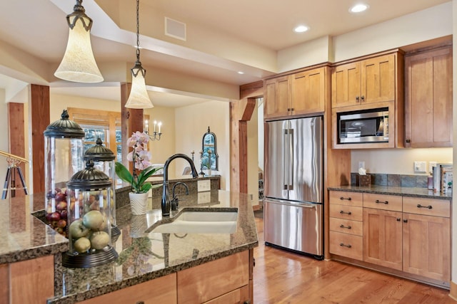 kitchen with appliances with stainless steel finishes, dark stone countertops, light hardwood / wood-style floors, sink, and hanging light fixtures