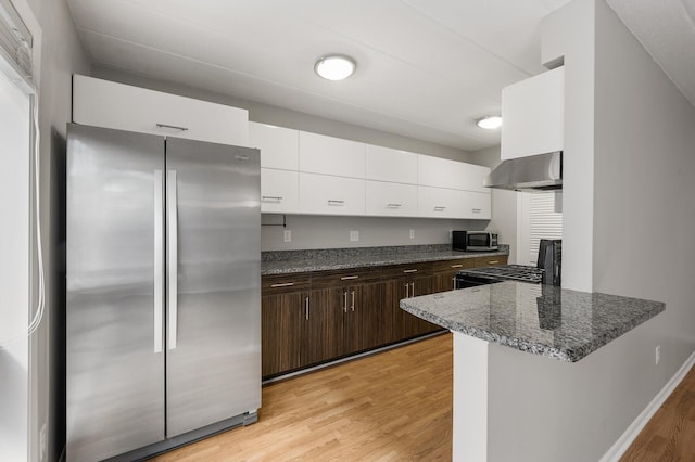 kitchen featuring white cabinets, wall chimney range hood, dark stone countertops, dark brown cabinets, and stainless steel appliances