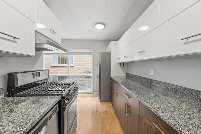 kitchen featuring light wood-type flooring, stainless steel appliances, dark stone countertops, white cabinets, and range hood