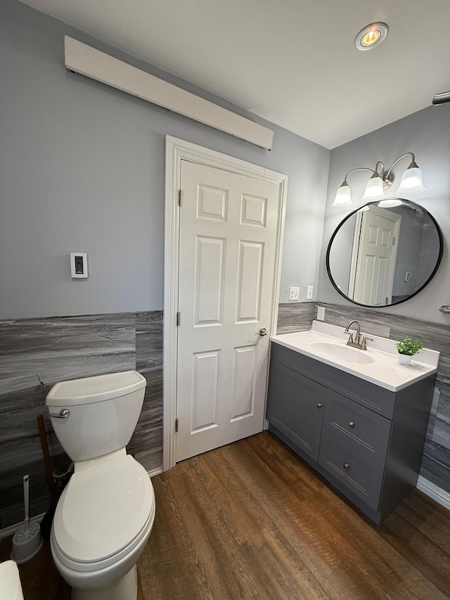 half bathroom featuring toilet, a wainscoted wall, wood finished floors, vanity, and tile walls