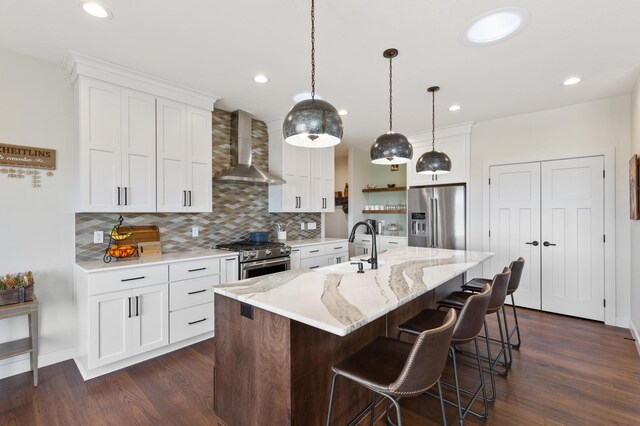 kitchen featuring decorative light fixtures, wall chimney exhaust hood, an island with sink, premium appliances, and white cabinetry