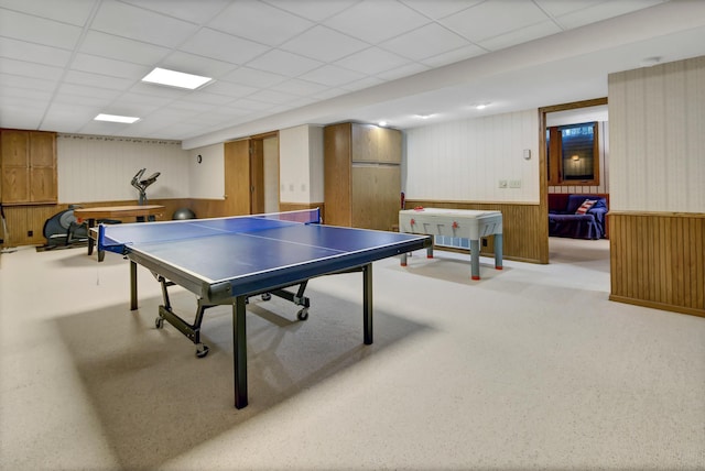 rec room with a paneled ceiling, wooden walls, and light colored carpet