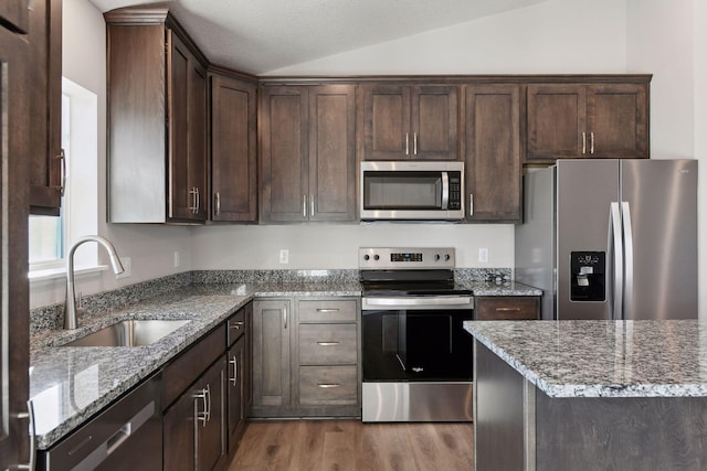 kitchen featuring lofted ceiling, sink, light stone countertops, appliances with stainless steel finishes, and dark brown cabinets