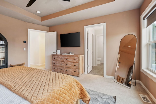 bedroom with ensuite bath, ceiling fan, a raised ceiling, light colored carpet, and multiple windows