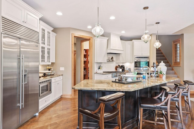 kitchen with premium range hood, built in appliances, decorative light fixtures, and white cabinets