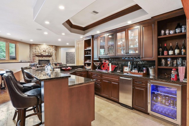 bar with wine cooler, a stone fireplace, dark brown cabinets, a raised ceiling, and dark stone counters