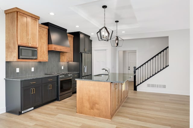 kitchen with premium range hood, pendant lighting, a tray ceiling, a center island with sink, and appliances with stainless steel finishes