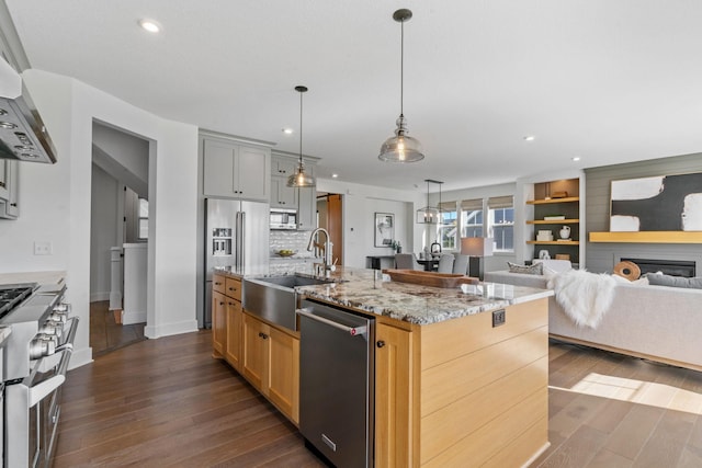 kitchen featuring wall chimney range hood, hanging light fixtures, stainless steel appliances, light stone counters, and a large island with sink