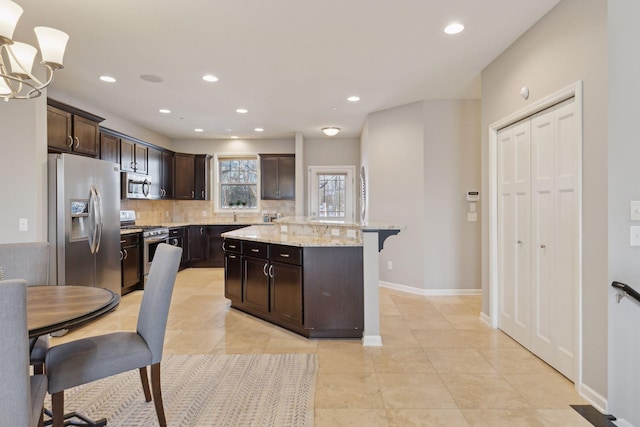 kitchen with decorative light fixtures, a kitchen island, light stone countertops, appliances with stainless steel finishes, and dark brown cabinets