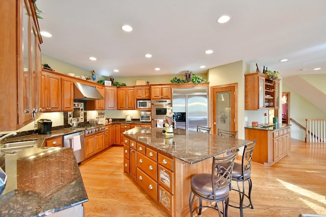 kitchen with sink, built in appliances, wall chimney exhaust hood, dark stone countertops, and a kitchen island