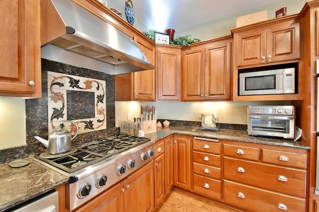 kitchen featuring decorative backsplash, dark stone counters, wall chimney exhaust hood, stainless steel appliances, and light hardwood / wood-style flooring