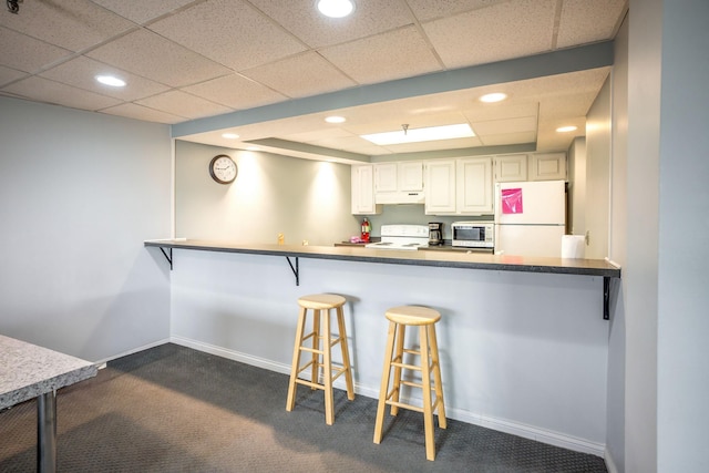 kitchen featuring kitchen peninsula, a paneled ceiling, dark carpet, white appliances, and white cabinetry