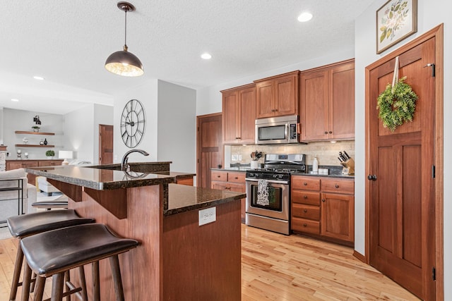 kitchen with backsplash, light hardwood / wood-style floors, pendant lighting, a kitchen island with sink, and appliances with stainless steel finishes