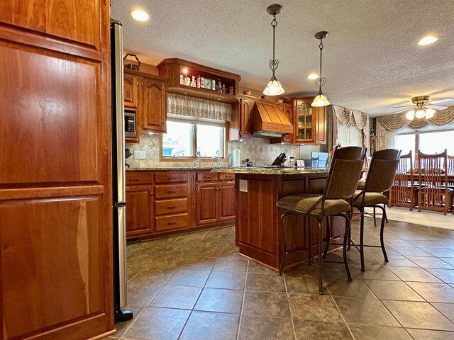 kitchen featuring a center island, hanging light fixtures, ceiling fan, light stone countertops, and custom range hood