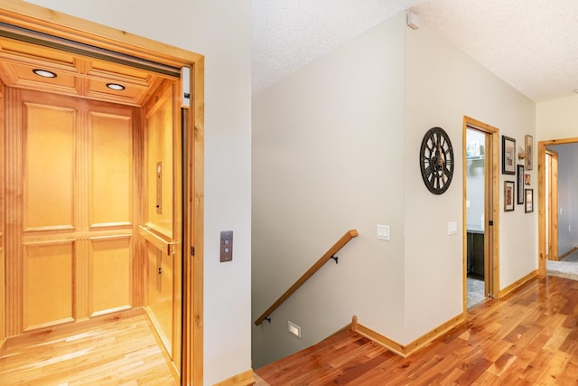 hallway with elevator, a textured ceiling, and light hardwood / wood-style flooring