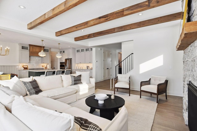 living room featuring beam ceiling, sink, an inviting chandelier, a stone fireplace, and light hardwood / wood-style flooring