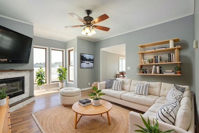 living area featuring baseboards, ceiling fan, wood finished floors, crown molding, and a stone fireplace