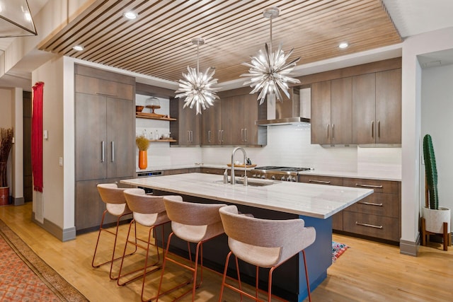 kitchen with wall chimney range hood, a center island with sink, sink, dark brown cabinetry, and a chandelier