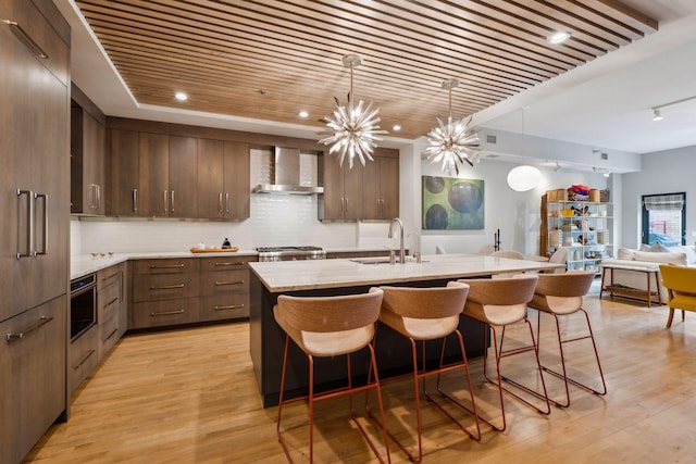 kitchen featuring wall chimney exhaust hood, sink, backsplash, a kitchen island with sink, and light wood-type flooring