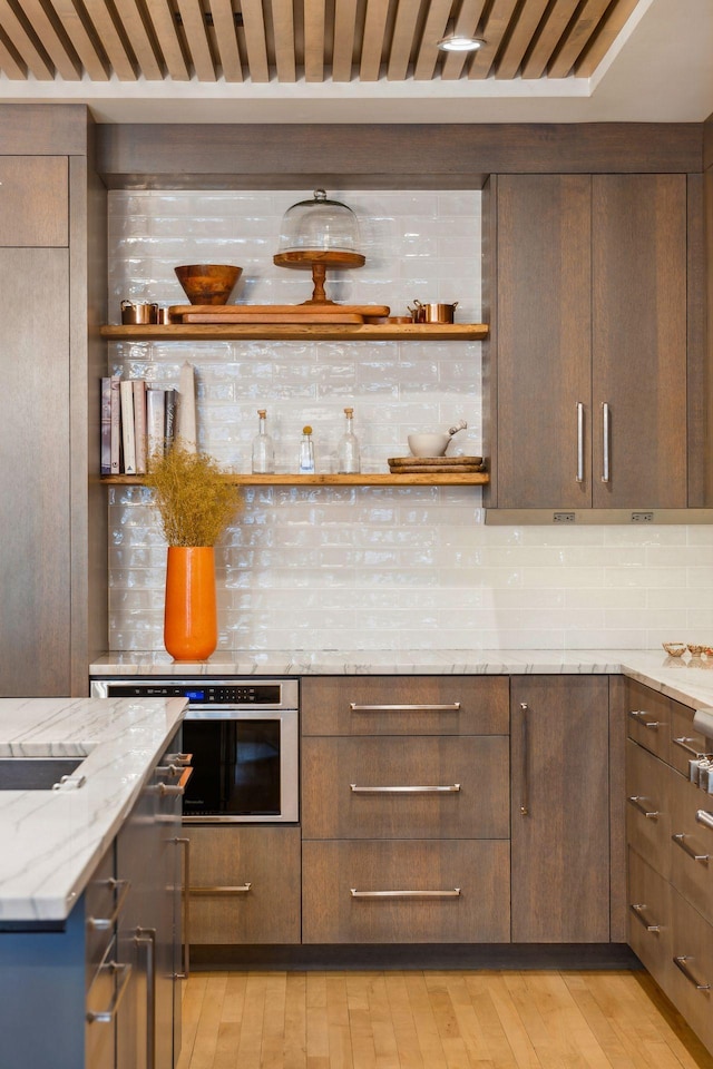 bar featuring oven, decorative backsplash, light hardwood / wood-style flooring, light stone countertops, and dark brown cabinets