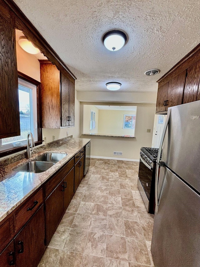 kitchen with appliances with stainless steel finishes, sink, light stone countertops, dark brown cabinets, and a textured ceiling