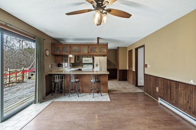 kitchen featuring kitchen peninsula, white appliances, a baseboard heating unit, a breakfast bar area, and wood walls