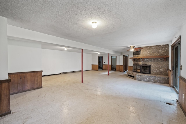 basement featuring ceiling fan, wooden walls, a baseboard heating unit, and a brick fireplace