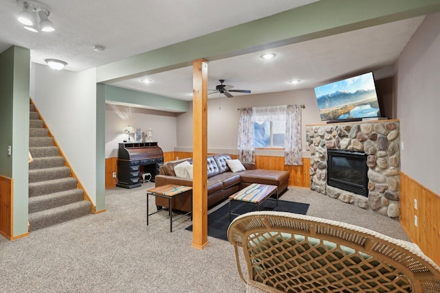 carpeted living room featuring ceiling fan, a stone fireplace, a textured ceiling, and wooden walls