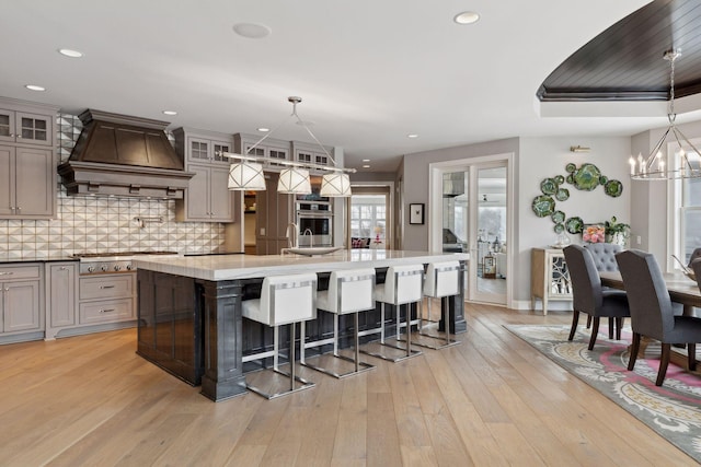 kitchen with custom exhaust hood, light hardwood / wood-style floors, stainless steel gas cooktop, a large island, and decorative light fixtures