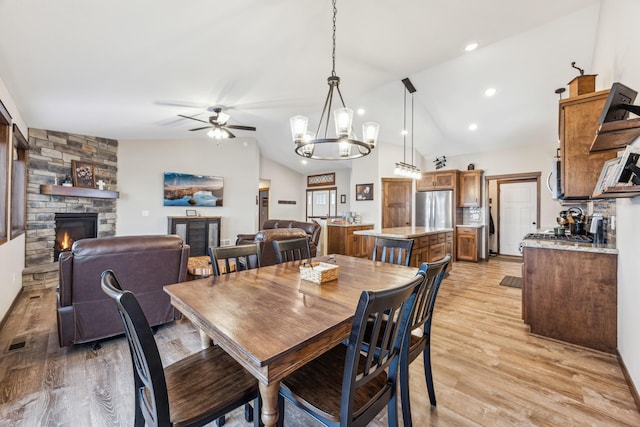 dining room with lofted ceiling, ceiling fan with notable chandelier, a stone fireplace, and light hardwood / wood-style flooring