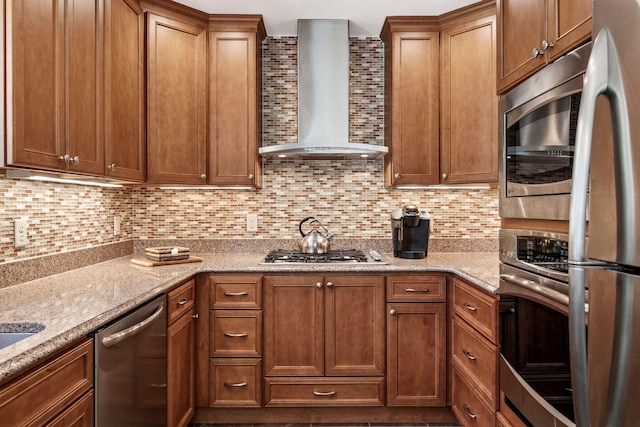 kitchen with wall chimney range hood, appliances with stainless steel finishes, tasteful backsplash, brown cabinetry, and light stone counters