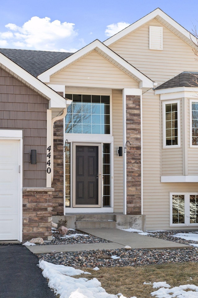 snow covered property entrance with stone siding, an attached garage, and roof with shingles