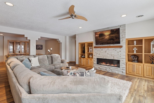 living room featuring built in shelves, a stone fireplace, a textured ceiling, ceiling fan, and hardwood / wood-style floors