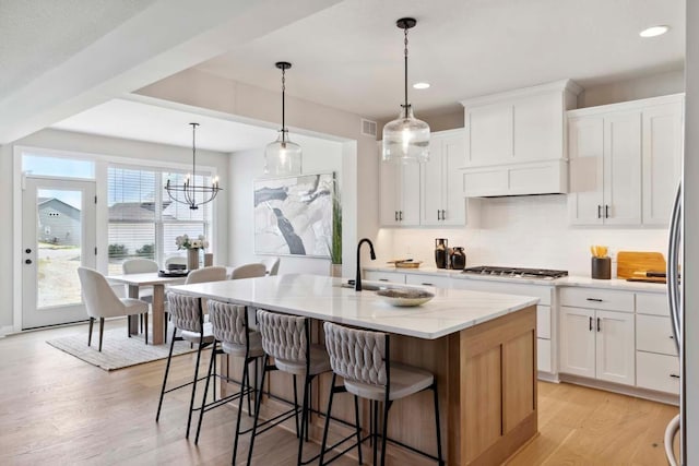 kitchen with white cabinetry, hanging light fixtures, an inviting chandelier, light hardwood / wood-style floors, and a kitchen island with sink