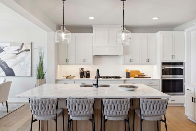 kitchen featuring a kitchen island with sink, white cabinets, sink, light stone countertops, and stainless steel double oven